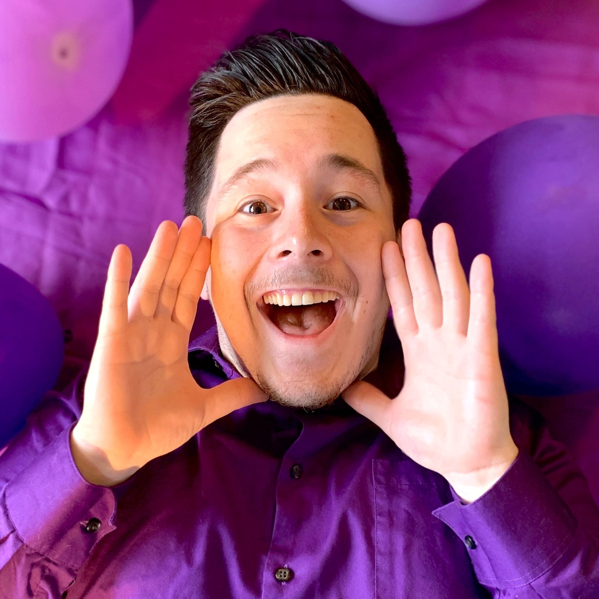 Smiling man in purple, surrounded by purple balloons, frames his face with his hands