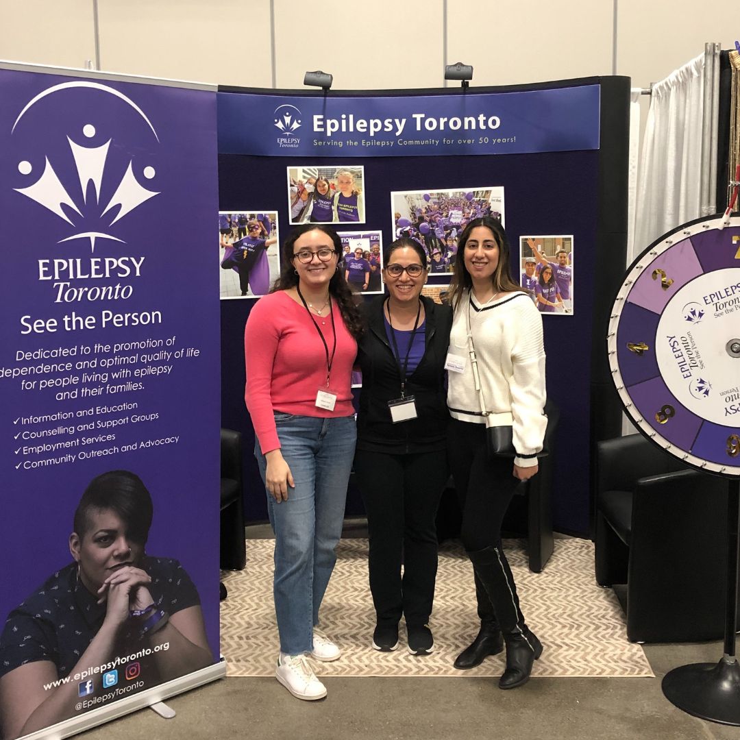 WomensShow2023 Three people wearing lanyards stand with their arms around each other smiling, in front of an Epilepsy Toronto display booth