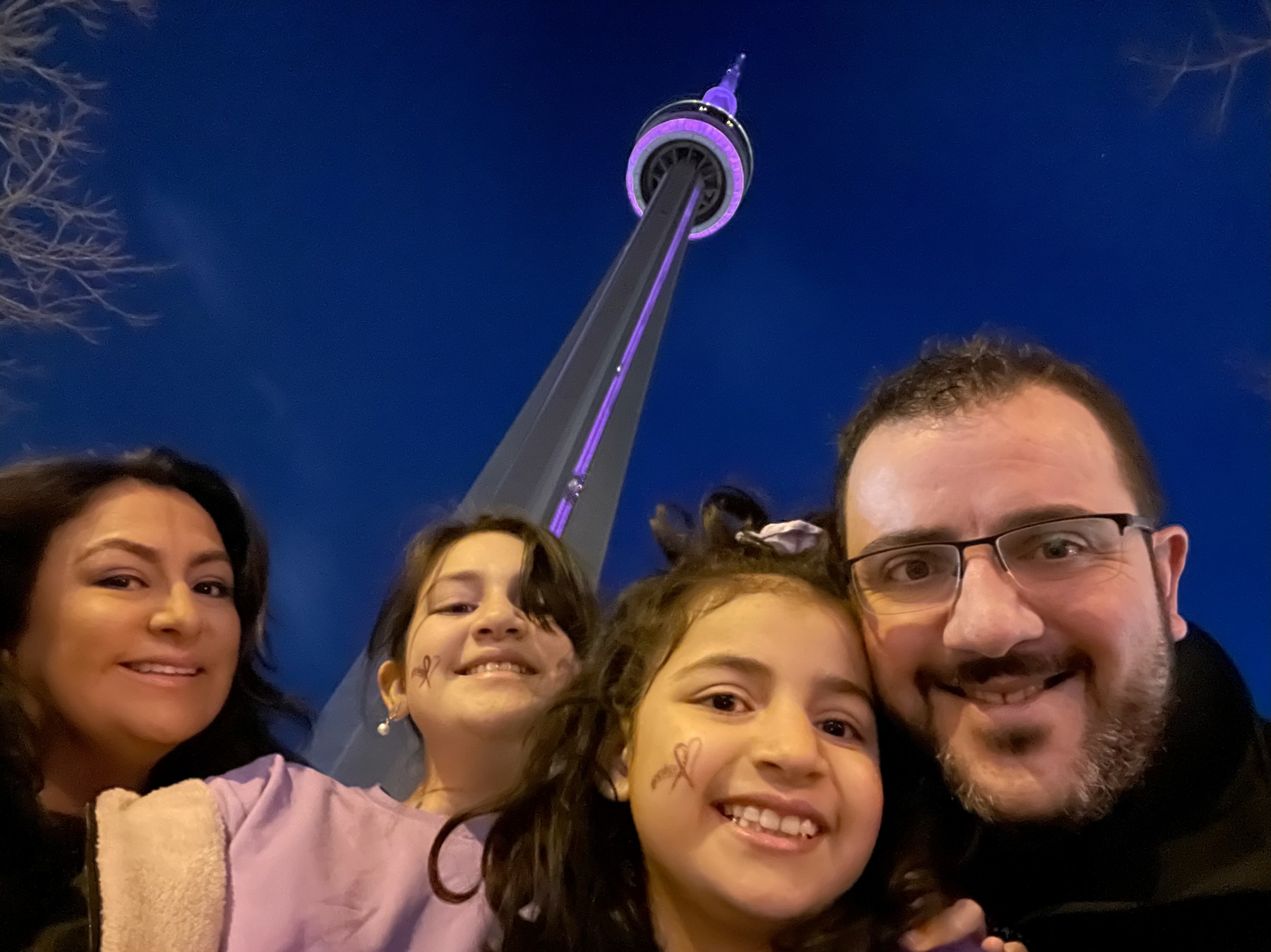 GrecoFamily(PurpleDay) Smiling family of 4 take selfie in front of the CN tower lit up purple.
