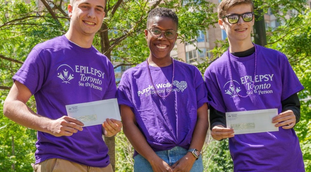 Three people wearing Epilepsy Toronto Purple Shirts stand smiling, the two on the ends each hold a certificate in front of them.