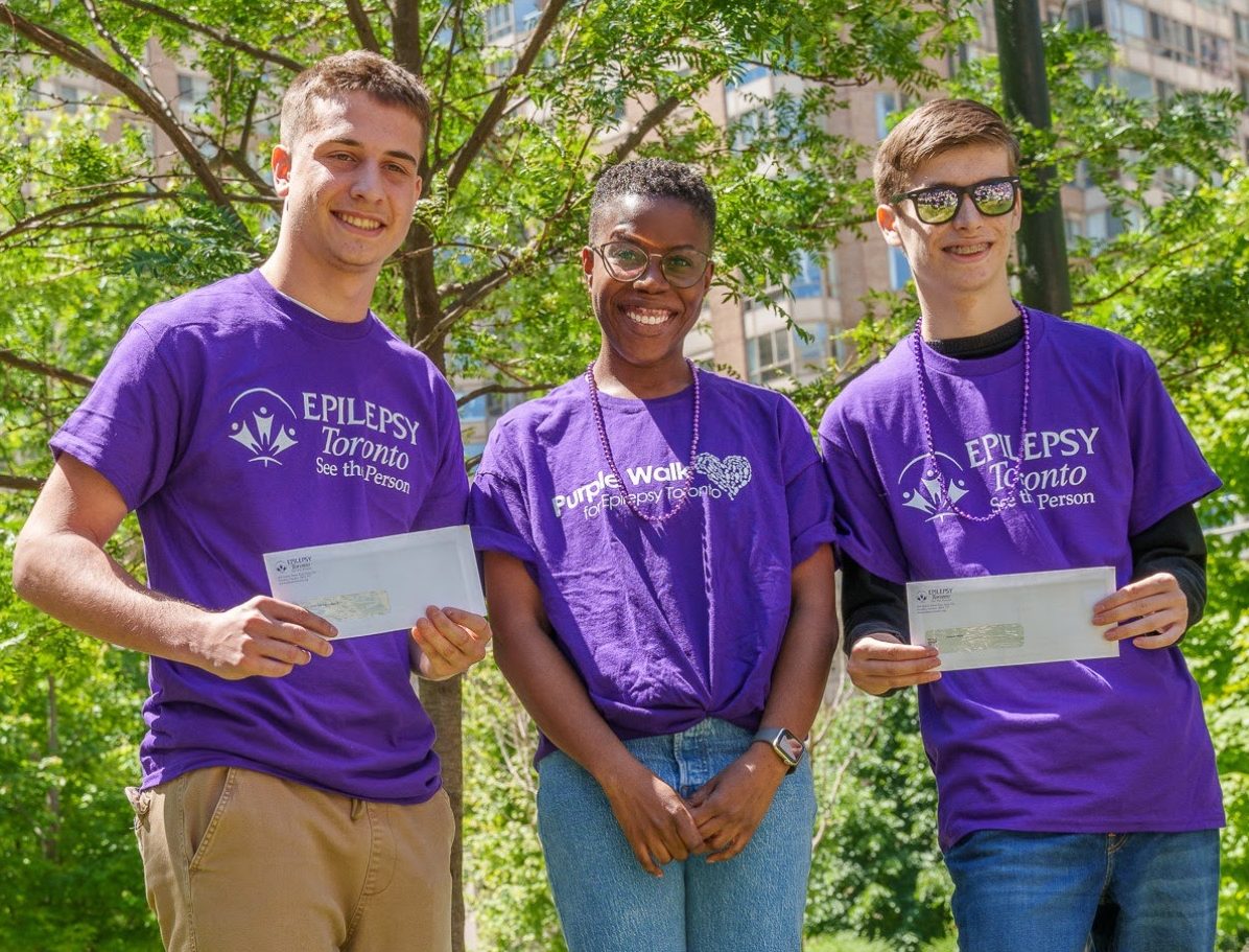 2024ScholarshipWinners Three people wearing Epilepsy Toronto Purple Shirts stand smiling, the two on the ends each hold a certificate in front of them.